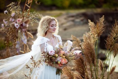 Gelin, gün batımında deniz kenarındaki kayalıklarda yapılan bir törende düğün süslemesinin yanında. Rüzgardan uçan peçe