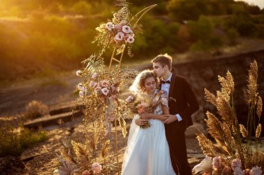 Gelin ve damat, gün batımında suyun yakınındaki bir kaya uçurumunda yapılan törende düğün dekorasyonunun yanında.