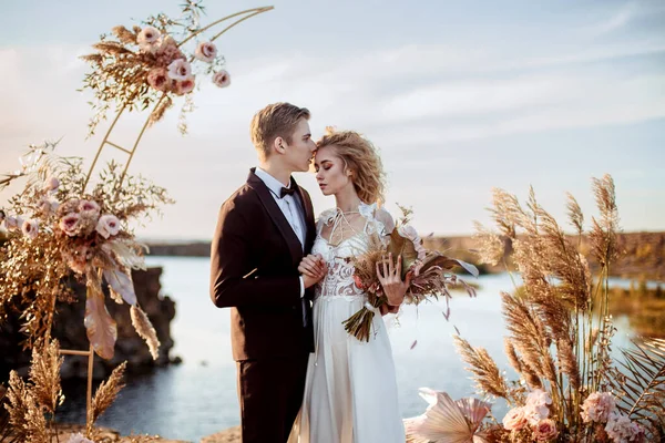 日没時に水の近くの岩の崖の上の儀式で結婚式の装飾の近くの花嫁と新郎 — ストック写真