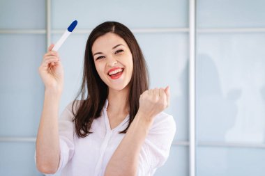 Genç bir kadın pozitif hamilelik testine bakarken seviniyor. Testi kamerada gösteriyor.