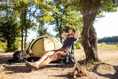 Bir adam ormandaki katlanır sandalyede çadırın yanında dinleniyor..
