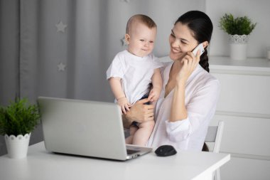 Genç anne, kucağında bir bebekle bilgisayarın başında çalışıyor.