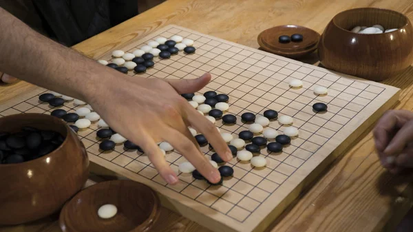 Mão jogando peças de pedra preto e branco no chinês Go ou Weiqi tabuleiro de jogo. Atividade interna com luz artificial . — Fotografia de Stock