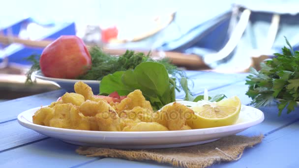 Жареные морепродукты с лимоном и рукколой на синем столе у моря и небольшие лодки в маленьком городке. Измир-Фока . — стоковое видео