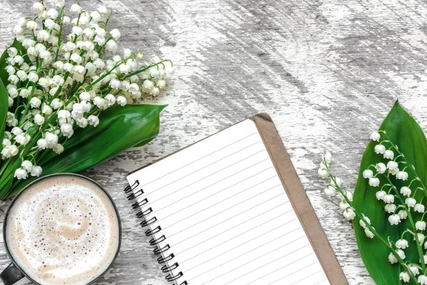 Φλυτζάνι καπουτσίνο με ανοιξιάτικα λουλούδια κρίνος της κοιλάδας και διαγραμμισμένο σημειωματάριο — Φωτογραφία Αρχείου