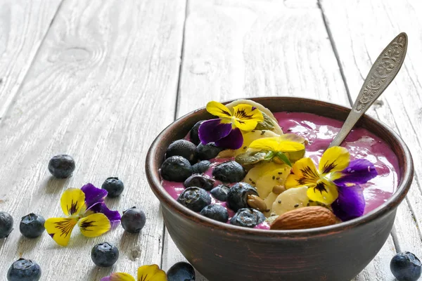 Ciotola di frullato con un cucchiaio condito con bacche fresche, banana, semi di chia, noci e fiori per una colazione sana — Foto Stock