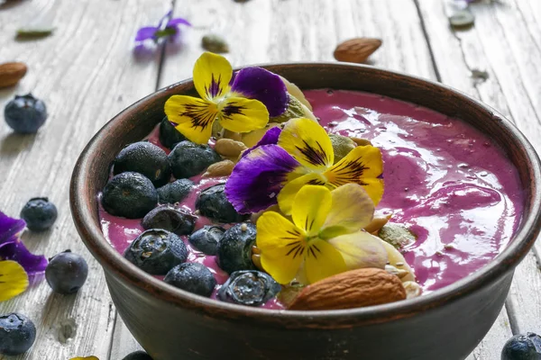 Ciotola di frullato di mirtilli condita con bacche fresche, banana, semi di chia e fiori per una sana colazione vegetariana . — Foto Stock