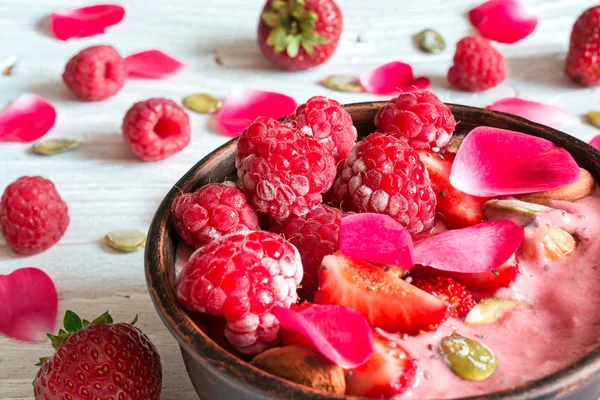 Hälsosam frukost. skål med röda bär smoothie med jordgubbar, hallon, nötter och frön dekorerad med rosa blomblad — Stockfoto