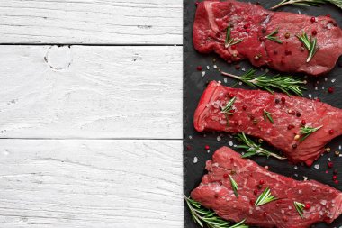 Çiğ et, sığır eti biftek baharat ve biberiye ahşap arka plan üzerinde siyah arduvaz kesme tahtası üzerinde. Üstten Görünüm ile