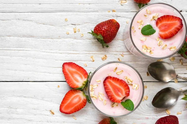 Yogur de fresa saludable con avena y menta en vasos con bayas frescas y cucharas sobre una mesa de madera blanca — Foto de Stock
