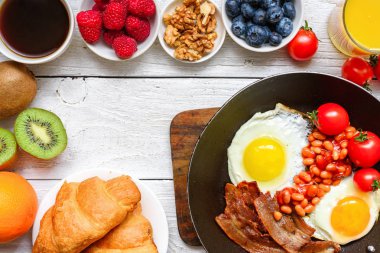 Kahvaltıda yumurta, domuz pastırması, fasulye, domates, kruvasan, meyve, fındık, kahve ve portakal suyu.