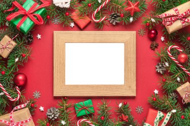 Çam ağacının dallarından, hediye kutularından, şekerlerden, kırmızı arka planda tatil süslemelerinden oluşan çerçevedeki noel kartı.