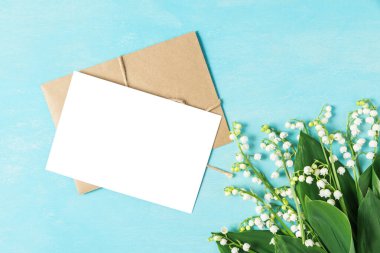 Boş tebrik kartı ve içinde bahar çiçekleri olan bir zarf. Mavi ahşap arka plandaki vadi zambağı. Model yap. Düz yat. Kopyalama alanı olan üst görünüm. düğün davetiyesi
