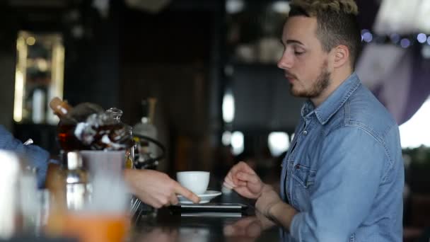 Un joven usa tabletas y bebe café mientras está sentado en el bar. — Vídeos de Stock