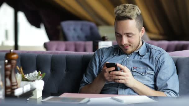 Hombre sentado en el restaurante, mensajes de texto en el teléfono móvil y sonriendo — Vídeos de Stock