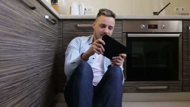 Joven jugando en la tableta sentado en la cocina en casa — Vídeos de Stock