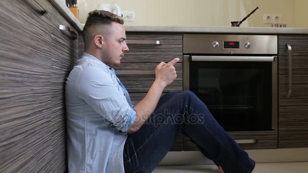 Hombre joven jugando en el teléfono inteligente sentado en la cocina en casa — Vídeos de Stock