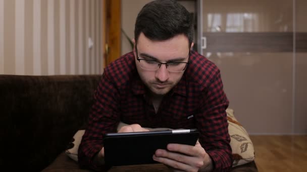 Junger Mann liegt auf dem Sofa und bekommt eine SMS auf einem Tablet — Stockvideo