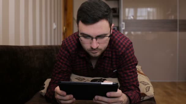 Jovem deitado no sofá e ganhando um sms em um tablet — Vídeo de Stock