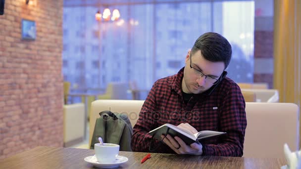 Volwassen man op telefoon praten en schrijven in een notitieblok in café — Stockvideo