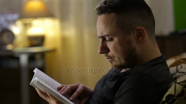 Homem lendo livro sentado no sofá à noite em casa — Vídeo de Stock