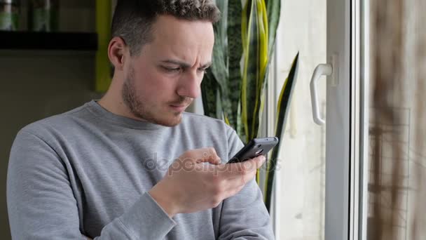 Jovem, homem feliz mensagens de texto no smartphone pela janela em casa — Vídeo de Stock