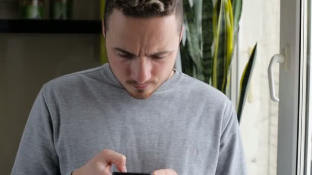 Молодой человек пишет смс на смартфоне, стоит у окна дома и смотрит в камеру. — стоковое видео