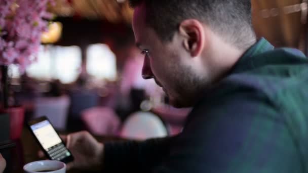 Joven sentado en un bar y enviando un mensaje en el teléfono móvil — Vídeos de Stock