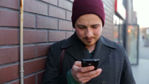 Jeune homme utilisant un smartphone debout près d'un mur de briques en ville — Video