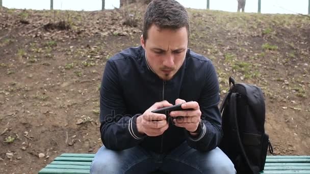 Молодий чоловік грає на сенсорному екрані телефону в парку — стокове відео