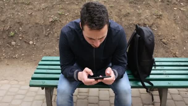 Молодой человек играет на сенсорном экране телефона в парке — стоковое видео