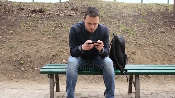 Fiatal férfi játszik a érint képernyő telefon a parkban — Stock videók