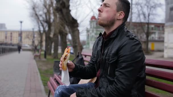 Seorang pria muda makan hotdog di kota di bangku cadangan — Stok Video