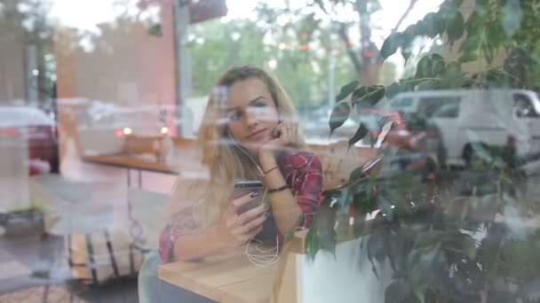 Girl listening to music in modern cafe . Street traffic is on window reflection — Stock Video