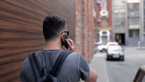 El tipo está caminando por la ciudad cerca de la pared de ladrillo y hablando por teléfono — Vídeo de stock