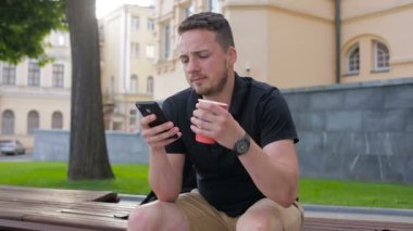 Genç bir adam kahve dışında şehir içme bir bankta oturmuş telefonu kullanın