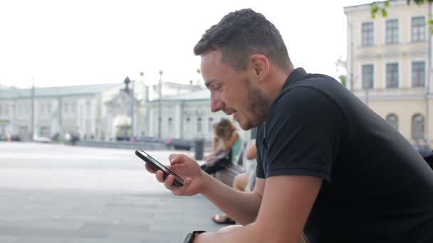Een jonge man gebruik telefoon zittend op een bankje buiten koffie drinken in de stad — Stockvideo