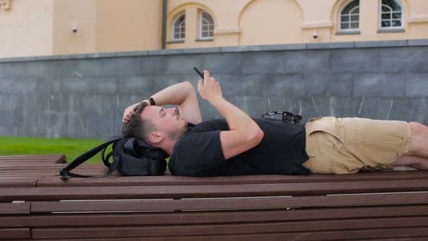 A young man uses a phone on a bench in the city — Stock Video