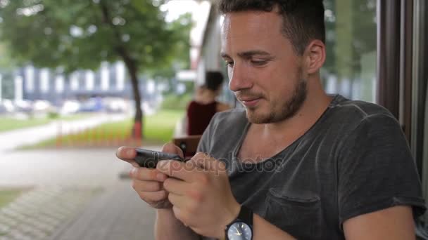 Ung man spelar på telefonen i ett café på gatan — Stockvideo