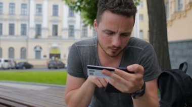 Adam parkta bir bankta akıllı telefon kullanarak internet üzerinden alışveriş sırasında kredi kartıyla