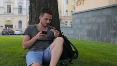 Adam bir ağacın altında oturup Park akıllı telefon kullanarak internet üzerinden alışveriş sırasında kredi kartıyla