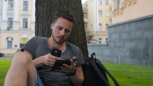 Człowiek z karty kredytowej podczas zakupów za pośrednictwem Internetu przy użyciu smartfona w parku siedzi pod drzewem — Wideo stockowe