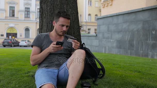 Pria dengan kartu kredit selama berbelanja melalui internet menggunakan telepon pintar di taman duduk di bawah pohon — Stok Video