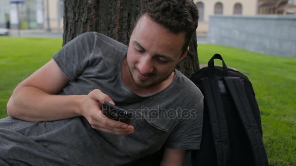 Der Kerl benutzt das Telefon unter einem Baum in einem geparkten unter einem Baum sitzend — Stockvideo