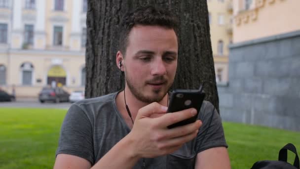 El tipo escucha música en el teléfono sentado debajo de un árbol en un aparcamiento debajo de un árbol — Vídeos de Stock