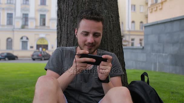 En kille spelar ett spel på telefonen medan du sitter under ett träd i en park som sitter under ett träd — Stockvideo