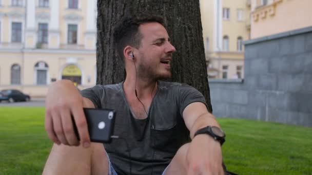 Ten chlap poslouchat hudbu na telefonu sedí pod stromem v zaparkovaných pod stromem — Stock video