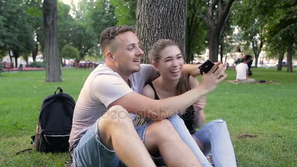 Guy with a girl doing selfie in a park under a tree — Stock Video