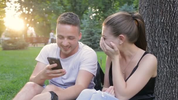 Guy mostra barzellette ragazza al telefono mentre seduto in un parco sotto un albero — Video Stock