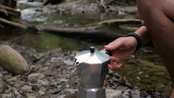 Um homem coloca uma cafeteira em um fogão em um acampamento na margem do rio — Vídeo de Stock